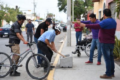 Grupo de ciudadanos y ciclistas en La Paz, Baja California Sur, rompiendo una banqueta de concreto que bloquea el paso peatonal. Una persona en silla de ruedas observa la acci&oacute;n junto a otros vecinos que se&ntilde;alan y participan con herramientas.