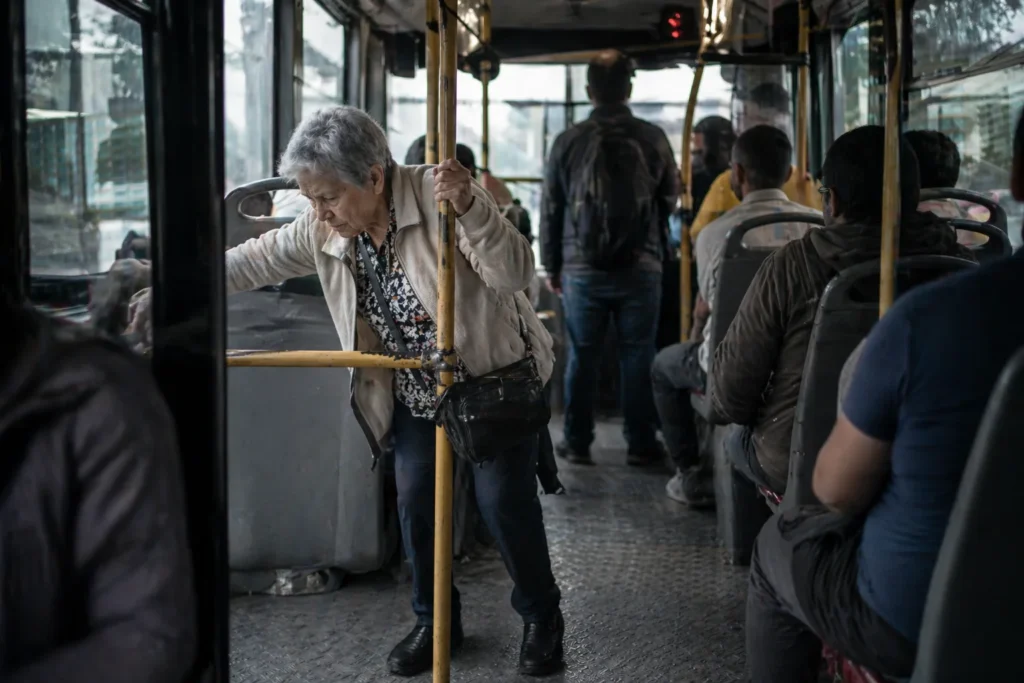 Persona adulta mayor intentando desplazarse dentro de unidad de transporte público con barras de apoyo insuficientes en Colombia.