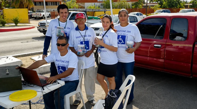 Grupo de voluntarios adultos participa en colecta comunitaria bajo carpa azul junto a camioneta roja, usando camisetas institucionales y recipientes para donativos.