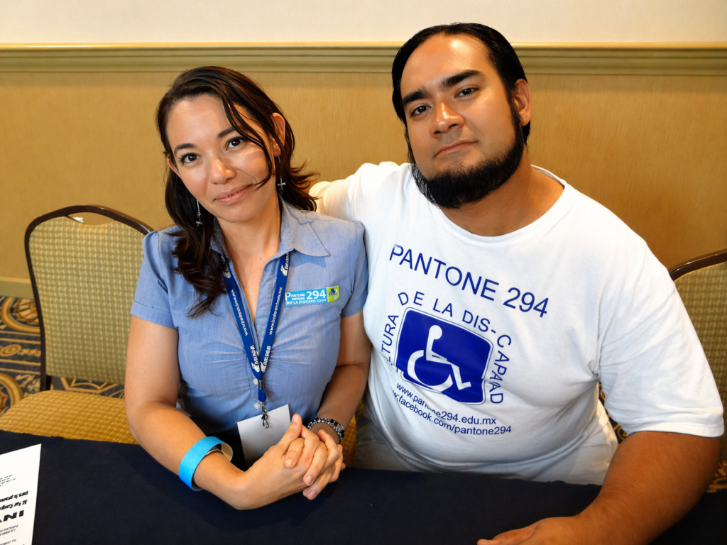 Dos personas adultas posan juntas en mesa de evento institucional, una con gafete y otra con camiseta de accesibilidad, en fotografía restaurada de alta calidad.