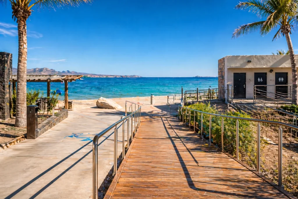 Pasarela de madera con rampas accesibles en la entrada de Playa El Coromuel en La Paz, con ba&ntilde;os adaptados frente al mar.