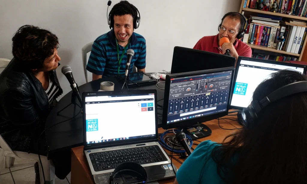 Equipo de locución participa en programa de radio o pódcast dentro de estudio improvisado, usando micrófonos, audífonos y computadoras durante una transmisión.