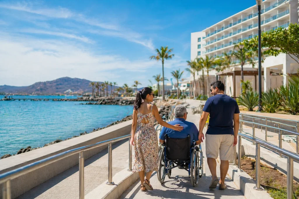 Malec&oacute;n de La Paz Baja California Sur con hotel accesible al fondo y familia acompa&ntilde;ando a persona mayor en silla de ruedas durante paseo junto al mar.