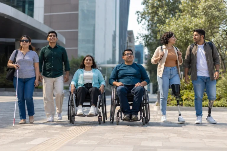 Grupo diverso de personas adultas con discapacidad desplazándose en plaza pública moderna de México, con usuarios de silla de ruedas, bastón blanco y prótesis visibles.