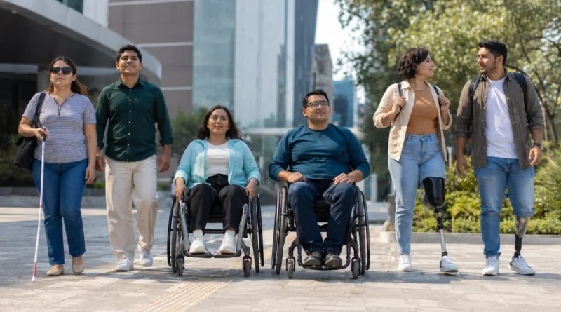 Grupo diverso de personas adultas con discapacidad desplazándose en plaza pública moderna de México, con usuarios de silla de ruedas, bastón blanco y prótesis visibles.